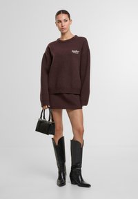 Dunkelroter Pullover mit gerippten Bündchen, kombiniert mit einem passenden Rock. Schwarze Handtasche mit silbernen Akzenten und kniehohe schwarze Stiefel mit spitzen Zehen.