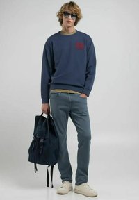 Felpa blu navy con logo rosso, camicia a strati verde chiaro, jeans azzurri e sneakers bianche. Zaino nero con patta e spallacci.