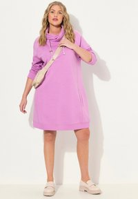 Vestido sudadera rosa con cuello de cisne y bolsillos laterales. La modelo lleva zapatos de plataforma beige y un bolso cruzado a juego.