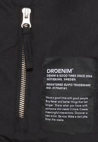 Tessuto nero con zipper argentato, caratterizzato da un'etichetta tessuta che riporta il nome del marchio "DRDENIM" e le istruzioni per la cura, oltre a un messaggio di sostenibilità.