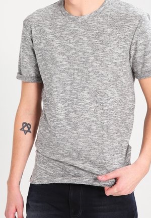 Kortærmet, meleret grå t-shirt med rund hals. Blød tekstur, tætsiddende design, og en tatovering synlig på venstre arm. Mørke jeans er iført.