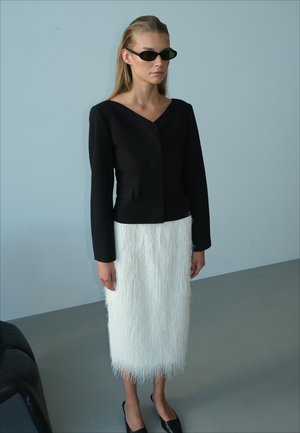 Femme aux cheveux blonds lisses porte des lunettes de soleil noires, un top noir ajusté à manches longues, une jupe midi blanche texturée et des chaussures noires pointues.