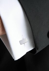 Bouton de manchette rectangulaire en argent fixé à une manche de chemise blanche texturée, avec un blazer foncé visible en arrière-plan.