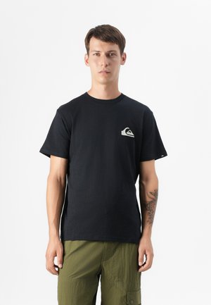 Ung mand i en sort T-shirt med et lille logo, iført olivengrønne cargo shorts, står foran en ensfarvet hvid baggrund.