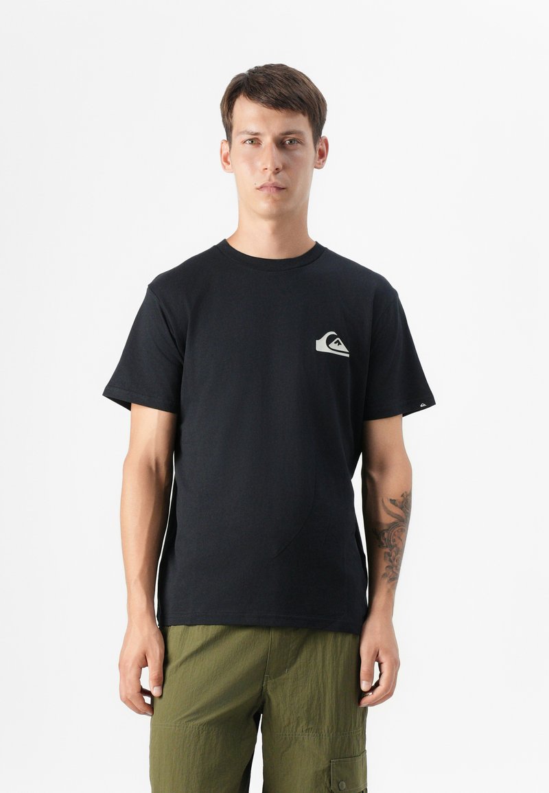 Jeune homme portant un t-shirt noir avec un petit logo, vêtu de shorts cargo verts olive, se tenant devant un fond blanc uni.