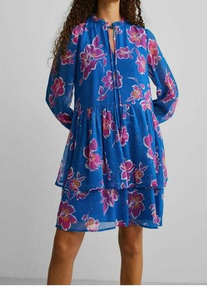 Femme portant une robe bleue à fleurs, manches longues, avec des fleurs roses et violettes et une jupe à volants, debout, les mains dans le dos.