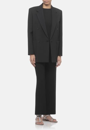 Persona che indossa un blazer doppiopetto nero, pantaloni neri a gamba larga e sandali piatti argentati borchiati, in piedi su sfondo bianco.