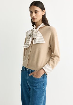 Frau mit langen dunklen Haaren, die einen beigen Cardigan, einen gestreiften Schleifenschal und blaue Jeans trägt, steht mit einer Hand in der Tasche.
