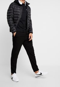 Mand iført sort puffjakke, sort polo-shirt, sorte bukser og hvide Nike sneakers, stående med en hånd i lommen mod en ensfarvet baggrund.