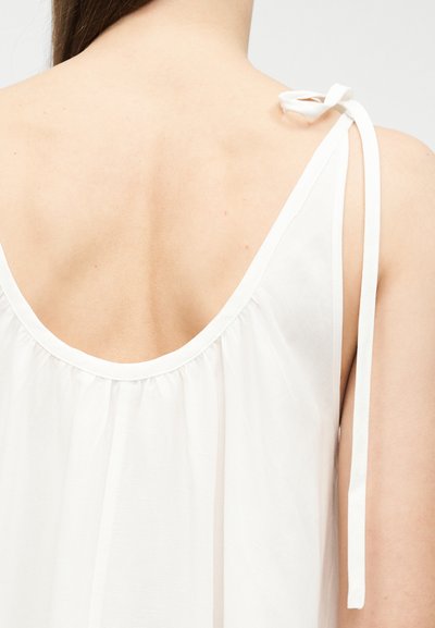 Κοντινό πλάνο του άνω μέρους της πλάτης ενός ατόμου που φοράει λευκή αμάνικη μπλούζα με δεμένο λουράκι στον δεξί ώμο.