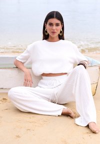 Witte gebreide crop top en wijdvallende broek, gemaakt van een textuurstof. Model zit op het zand nabij een strand, met grote ronde oorbellen en een ontspannen houding.