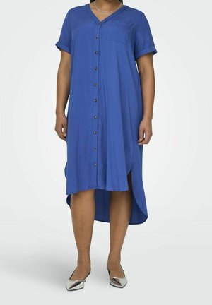 Kobieta w niebieskiej sukience z guzikami sięgającej do kolan, z krótkimi rękawami i srebrnymi punktowanymi balerinami, stoi na jednolitym tle.