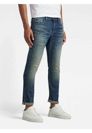 Mand iført oprullede, falmede blå jeans og hvide sneakers står foran en ensfarvet baggrund.