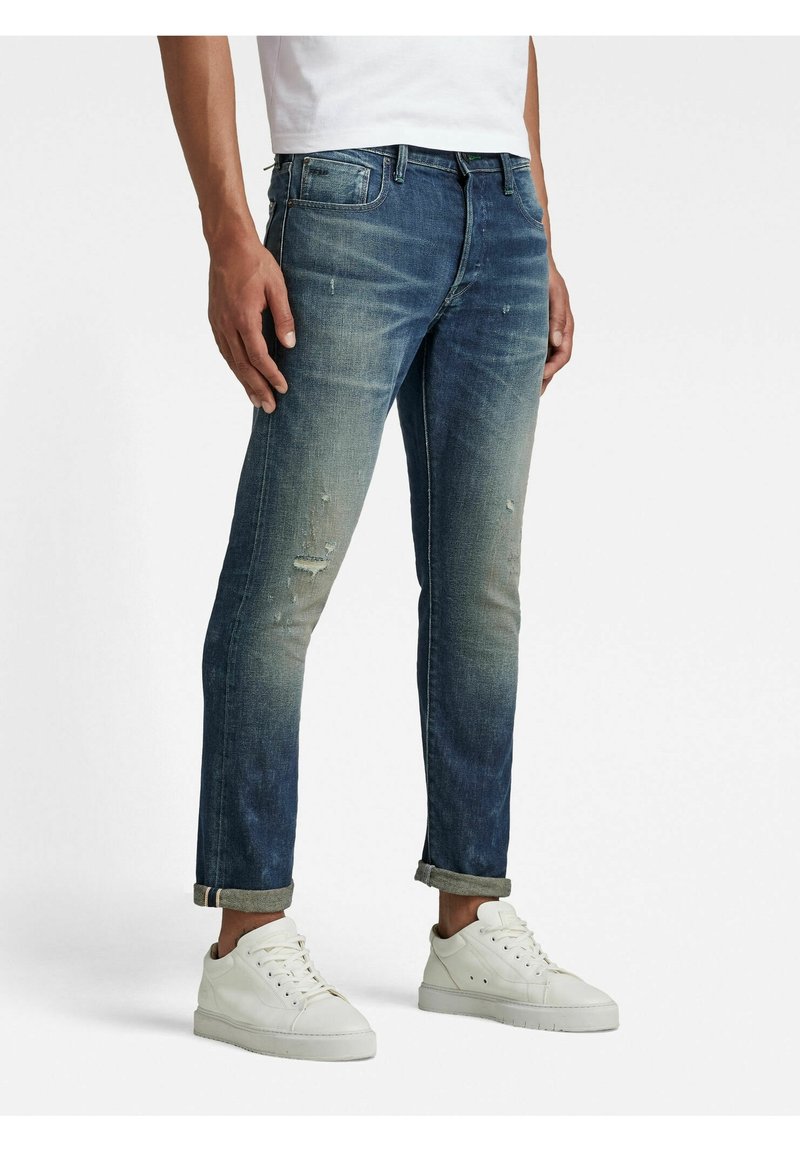 Homme portant un jean bleu délavé retroussé et des baskets blanches, debout devant un fond uni.