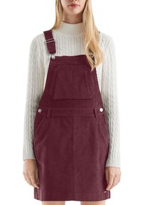 Burgunderfarbene Corduroy-Latzkleid mit verstellbaren Trägern, vorderer Tasche und seitlichen Taschen, getragen über einem cremfarbenen gerippten Rollkragenpullover.