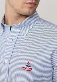 Camisa abotonada en rayas azul claro y blancas. Cuenta con un cuello clásico, logo bordado en rojo y azul, y botones blancos.