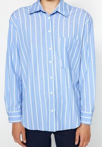 Ljusblå skjorta med knappar och vita vertikala ränder, långa ärmar, en bröstficka och rundad nederkant. Tillverkad av ett mjukt tyg.