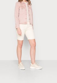 Lichtroze spijkerjack over een gestreept topje, gecombineerd met crème-kleurige bermudashorts en beige sneakers, geplaatst tegen een neutrale achtergrond.