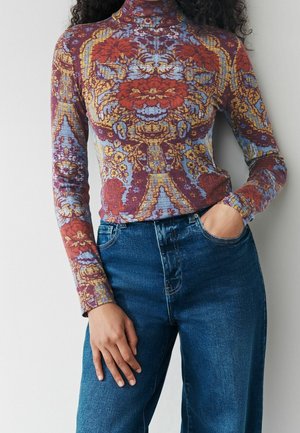 Femme portant un col roulé moulant à manches longues avec un motif floral multicolore et un jean bleu taille haute, une main dans la poche.