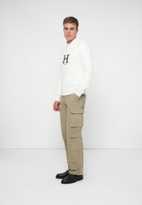 Felpa bianca con un grande logo nero "H", abbinata a pantaloni cargo beige con numerose tasche e scarpe scure.