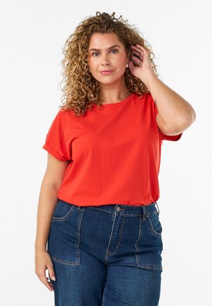 Kvinde med krøllet hår iført en skrigende rød kortærmet skjorte og blå jeans med høj talje, der rører ved sit øre med den ene hånd.