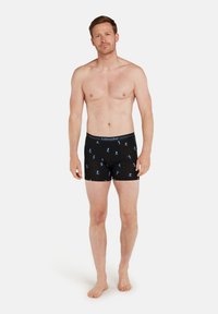 Bar överkropp vuxen man som står barfota iklädd svarta boxerkalsonger med blått skidmönster, mot en enkel vit bakgrund.