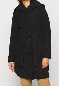 Svart, texturerad kappa med huva och långa ärmar. Har ett band i midjan och en lös passform, tillverkad av ett plyschigt, mjukt material.