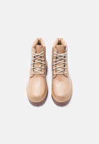 Γυαλιστερά μποτάκια από μπεζ δέρμα με κορδόνια και χοντρές καφέ σόλες, εμφανιζόμενα από πάνω σε λευκό φόντο.