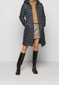 Mørkeblå dunjakke med hætte, lang længde, quiltet tekstur og sidelommer. Båret over en beige rullekrave og ternet nederdel, kombineret med sorte knæhøje støvler.