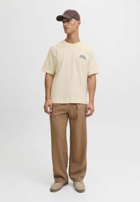 Cămașă din bumbac crem cu text verde "APPAREL STUDIOS", asortată cu pantaloni largi maro și pantofi slip-on bej. Casca maro.
