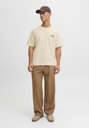 Creme bomulds t-shirt med grøn "APPAREL STUDIOS" tekst, parret med brune brede bukser og beige slip-on sko. Brun kasket.