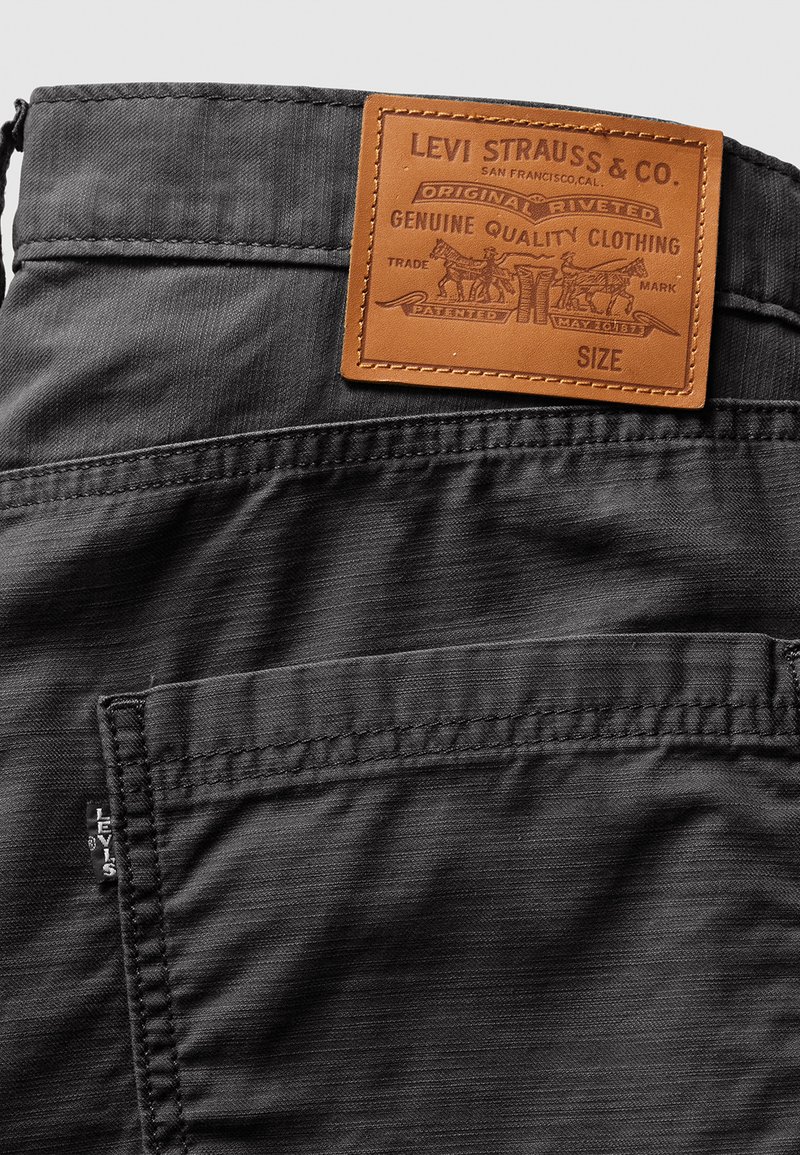 Pantalones de tela gris con un bolsillo de parche y una etiqueta de cuero marrón que detalla la información de la marca y del producto. Material texturizado con costuras visibles.