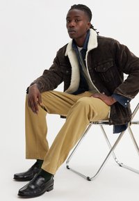 Jachetă din corduroi maro cu guler alb din blăniță, stratificată peste o cămașă din denim, asociată cu pantaloni khaki și mocasini negri, așezată pe un scaun cromat.