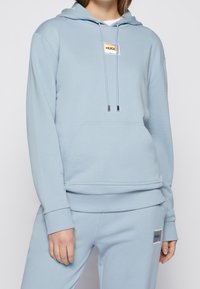 Sudadera azul claro hecha de un tejido suave, con un bolsillo canguro, cuerdas ajustables y un parche de logo holográfico en el pecho.