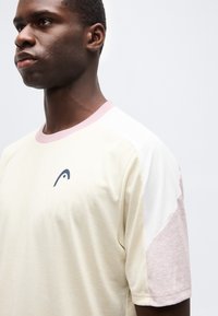 Νεαρός άνδρας που φοράει μπλουζάκι με κοντά μανίκια σε μπεζ, άσπρο και ανοιχτό ροζ χρώμα με ένα μικρό σκούρο λογότυπο στο στήθος, κοιτάζοντας προς τα αριστερά.