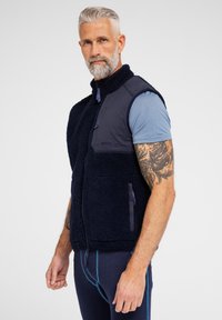 Marineblå fleece vest med en glat marineblå stof foran, lynlåslommer og en struktureret krop. Har kontrasterende lilla detaljer.
