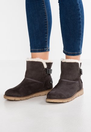 Bottes de cheville en daim marron avec une douce doublure en fourrure crème, une sangle décorative et un bouton rond, montées sur une semelle en caoutchouc texturée couleur tan.