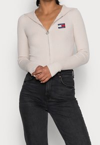 Light beige ribbed zip-up top with a hood and a small embroidered logo on the chest, paired with dark denim jeans.