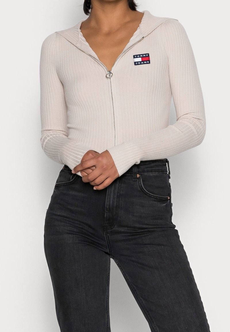 Light beige ribbed zip-up top with a hood and a small embroidered logo on the chest, paired with dark denim jeans.