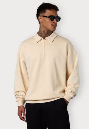 Beige, langärmliges Poloshirt mit Reißverschlusskragen, das einen lässigen Schnitt und ein gesticktes Logo aufweist, kombiniert mit schwarzen Hosen und Sonnenbrille.