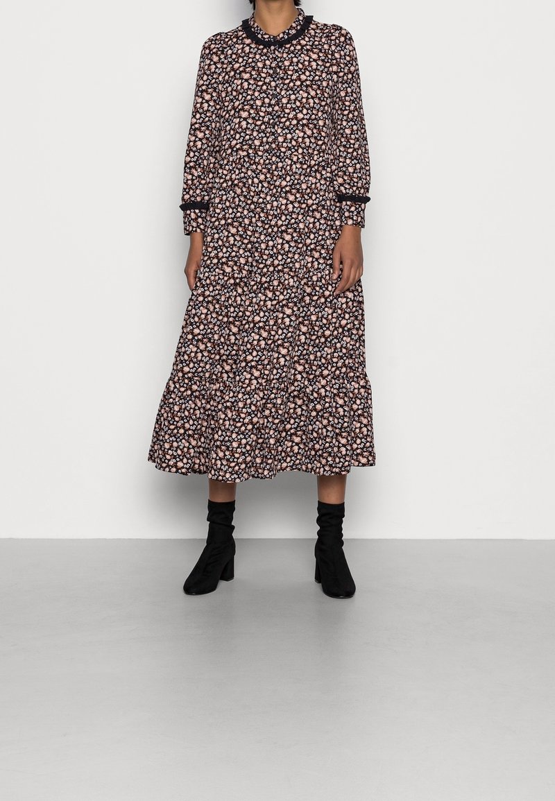 Femme portant une robe midi à manches longues à motif floral, associée à des bottines noires à talons, debout devant un fond uni.