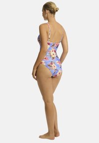 Model in een bloemen badpak, van achter gezien, met een lichtblauwe achtergrond die de levendige kleuren en het design van het badpak benadrukt.