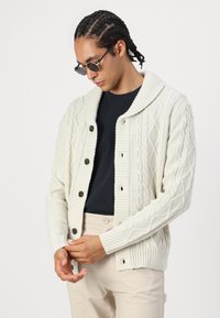 Geknoopte cardigan in gebroken wit, met een sjawlkol en ribgebreide manchetten, en bruine knopen. Draagt over een zwarte shirt, met een gladde textuur.