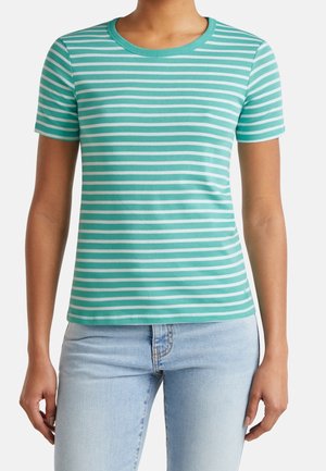 Kvinde iført en kortærmet, turkis og hvide horisontale stribet T-shirt kombineret med lyseblå jeans, stående mod en ensfarvet baggrund.