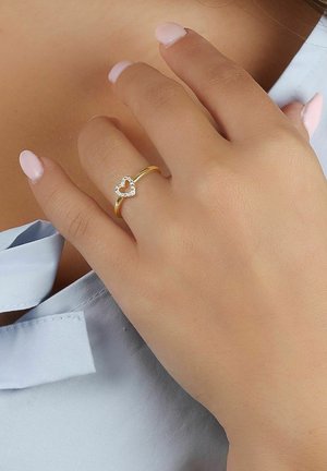 Guld ring med ett hjärtformat mönster accentuerat av klara stenar. Handen har en jämn textur, med ljusrosa nagellack.