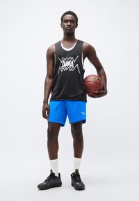 Μαύρη αμάνικη φανέλα μπάσκετ με λευκό γραφικό, μπλε σορτς, λευκές κάλτσες και μαύρα αθλητικά παπούτσια, κρατώντας μια μπάλα μπάσκετ.