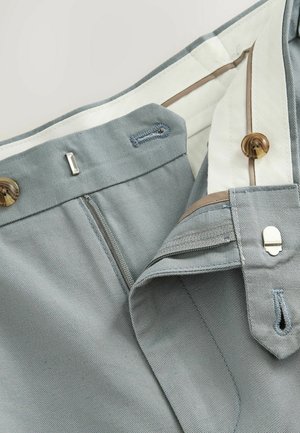 Svijetlosivi hlače s patentnim zatvaračem, smeđim gumbom i bijelim unutarnjim podstavom. Sadrže šivene detalje i metalnu kuku za zatvaranje.