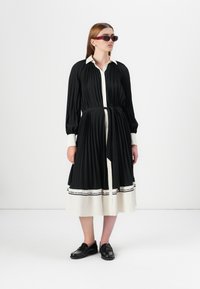 A model stands wearing a black pleated dress with a white collar, cinched at the waist, paired with dark sunglasses and black shoes.