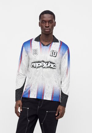 Homme portant un maillot de football à manches longues blanc, bleu et rouge avec le texte "replay inc." et le numéro 10, debout devant un fond blanc uni.