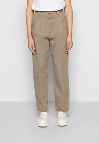 Beige high-waisted broek met plooien, een relaxte pasvorm en riemlussen, gecombineerd met witte platform sneakers. De stof lijkt soepel.